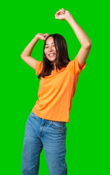 A young woman is expressing happiness by dancing with her arms raised in a lively manner. She wears an orange shirt and blue jeans, exuding a carefree vibe in a playful scene.