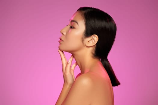 A young woman with a smooth complexion poses elegantly in profile, her hand gently touching her chin. The vibrant pink background enhances her graceful features.
