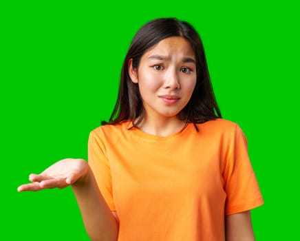A young woman wearing an orange shirt stands with a puzzled expression, raising one hand in a questioning gesture. The vibrant green background enhances her expression of confusion.