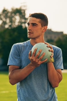 A young man stands on a field holding a shot put, focused and ready to throw. The warm sunlight highlights his concentration while training outdoors.