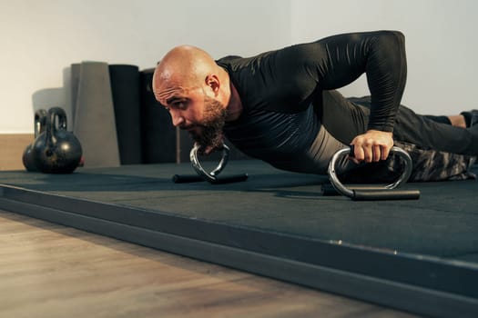 A man in a fitted long-sleeve shirt exercises by doing push-ups on gym mats. He uses workout equipment to enhance his strength training routine. The gym features a clean and modern design.