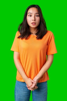A young woman with long dark hair wears an orange shirt, displaying a surprised look while standing with her hands clasped in front of her, surrounded by a vibrant green background.