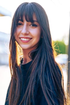 A youthful woman with long hair and bangs is smiling surrounded by soft sunlight. The background features urban elements, creating a vibrant atmosphere.