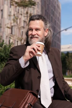 happy successful mature male in elegant clothes drinking a cup of takeaway coffee sitting on urban square, concept of business and middle age man lifestyle