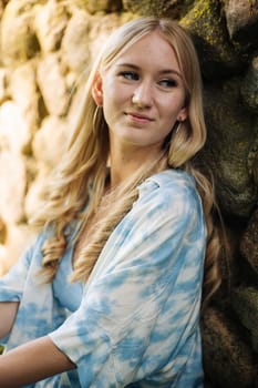 Young woman with blond hair and blue tie dye shirt sitting against an old stone wall, enjoying a moment of peace