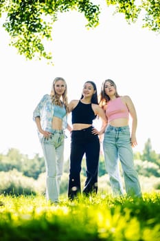 Three young women, school friends, are posing together in a park during their summer holidays, enjoying the sunny weather and green landscape