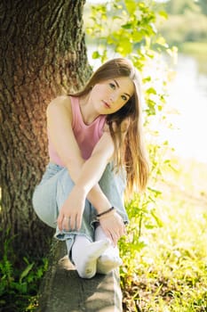 Young woman in casual attire relaxing under a tree by the lake, savoring the warmth of a summer day and embracing nature's tranquility
