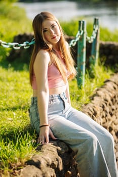 Young woman sitting on a low stone wall by a serene river in a park, soaking up the warm summer sun and enjoying a peaceful moment