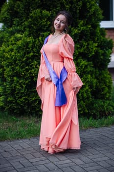 Graduate student celebrating graduation in a peach dress and sash, radiating happiness and pride in a lush garden setting