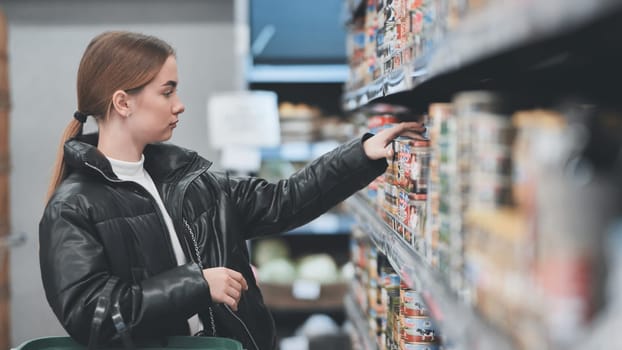 Young woman shopping groceries, reaching for a can on a supermarket shelf, making purchasing decisions in a retail environment