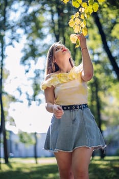 Fashionable young woman gently touching colorful autumn leaves on a tree branch in a sunny park, enjoying the fall season