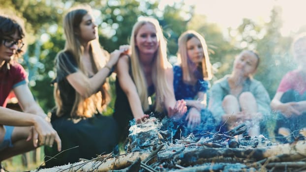 Group of school friends warming up around dying campfire during summer camping trip in a forest