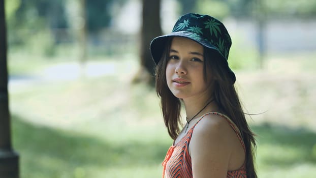 Portrait of a teenage girl wearing a bucket hat featuring a marijuana leaf design, posing in a park during a sunny summer day
