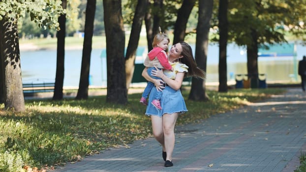 Young mother holding her daughter while walking in a park near a lake during a sunny summer day