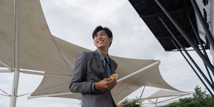 A smiling businessman dressed in a suit, holding a sandwich outdoors under a large fabric canopy, reflecting a joyful moment during a break.