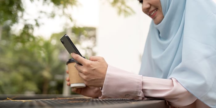 A Muslim businesswoman in hijab smiles as she uses her smartphone and holds a coffee cup, enjoying a relaxed moment in an outdoor setting.
