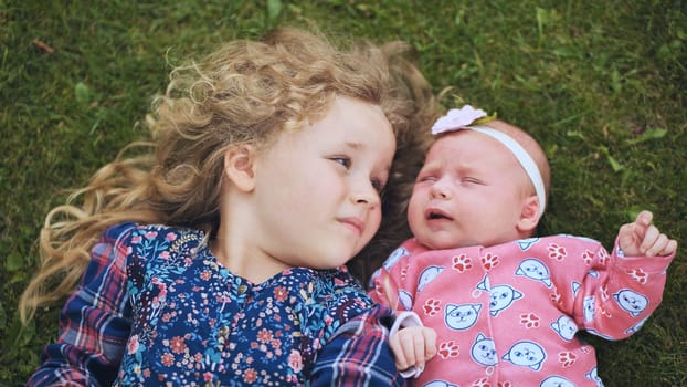 Newborn and toddler sisters relaxing on grassy lawn, sharing tender moment during sunny family gathering, radiating pure affection and happiness