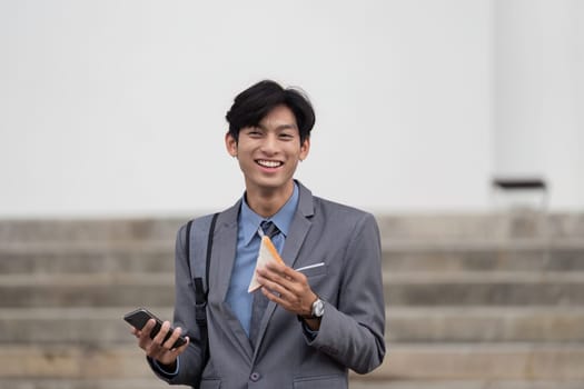 A young businessman dressed in a suit, smiling while holding a smartphone and sandwich outdoors near a staircase, reflecting a casual lunch break.