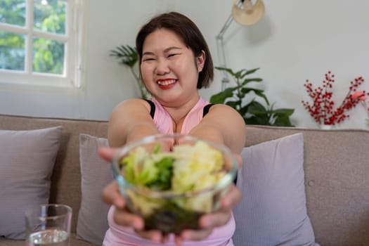 An overweight woman smiling and presenting a fresh salad, encouraging healthy eating habits and weight loss in a welcoming home setting.