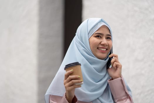 A Muslim businesswoman smiling while speaking on the phone and holding a coffee cup, highlighting professionalism and positivity in a casual setting.