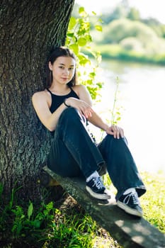 Serene moment of peace as a young woman enjoys the tranquility of nature, sitting on a wooden plank beneath a large tree by a river