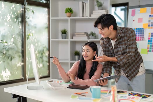 A diverse graphic design team happily collaborating and presenting ideas while using a computer and tablet in a bright, creative workspace.
