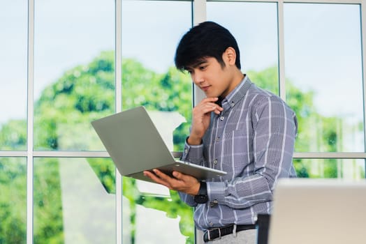 Asian businessman smiling and working on a laptop computer stand near window in office. Portrait of handsome man standing and online meeting with computer notebook in office lobby