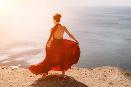 Side view a Young beautiful sensual woman in a red long dress posing on a rock high above the sea during sunrise. Girl on the nature on blue sky background. Fashion photo.