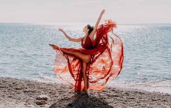 Side view a Young beautiful sensual woman in a red long dress posing on a rock high above the sea during sunrise. Girl on the nature on blue sky background. Fashion photo.