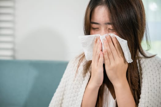 Sick female sitting under blanket on sofa and sneeze with tissue paper in living room, fever caught cold, Asian young woman she sick blowing nose sneezing in tissue at home, medicine healthcare