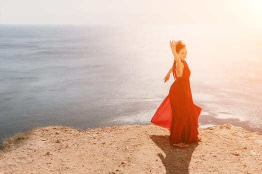 Side view a Young beautiful sensual woman in a red long dress posing on a rock high above the sea during sunrise. Girl on the nature on blue sky background. Fashion photo.