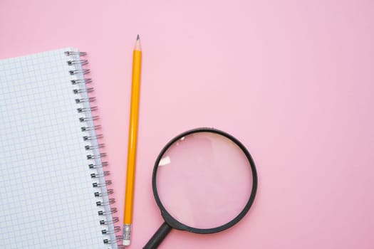 Magnifying glass, pencils and a blank white notepad on a spring on a pink background. Back to school concept. Copy space for text