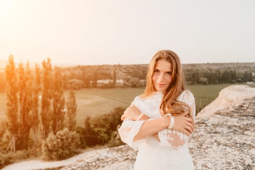 Romantic beautiful bride in white dress posing with sea and mountains in background. Stylish bride standing back on beautiful landscape of sea and mountains on sunset