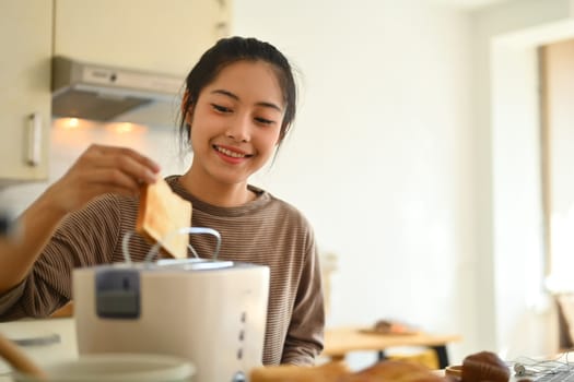 Beautiful young woman preparing breakfast, toasting bread with toaster in the kitchen. People, food and domestic life concept.