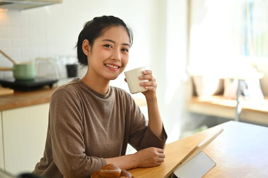 Positive asian woman drinking coffee and using digital tablet at kitchen counter in the morning.