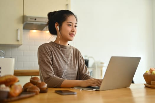 Millennial asian woman working online or browsing internet on laptop at kitchen counter.