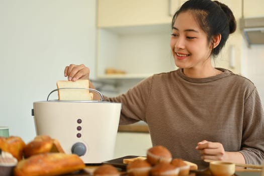 Happy young woman toasting bread with toaster in the kitchen. People, food and domestic life concept.