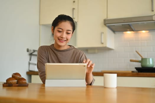 Smiling millennial asian woman drinking coffee and using digital tablet at kitchen counter in the morning.