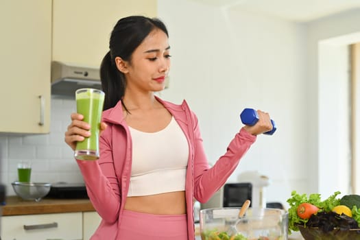 Sporty woman holding glass of fresh green vegetable juice and lifting dumbbell in kitchen. Healthy lifestyle concept.