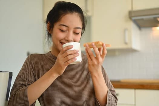 Relaxed young woman holding toast with jam and drinking coffee. People, food and domestic life concept.