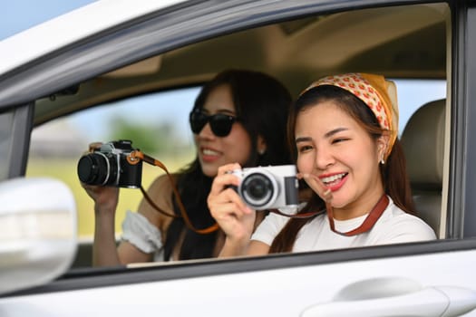 Two excited female tourists taking photos from a car window with vintage retro cameras during their summer road trip.