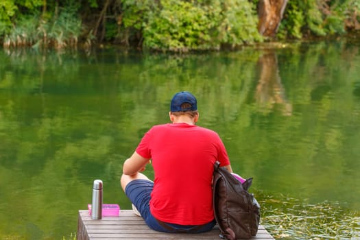 Man sitting back. An adult man resting in shade on shores lake was bleeding on beautiful sunny spring day. With a thermos and breakfast in a red t-shirt and shorts