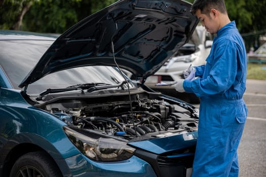 Close-up of an Asian mechanic's hand in a blue shirt, writing repair notes on a paper while working on a car in an auto repair shop.