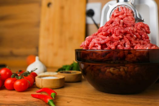 Side view on a wooden background: dark dishes with red minced meat inside, which comes out of a metal meat grinder, spices, herbs and vegetables are nearby. The concept of healthy eating, cooking. High quality photo