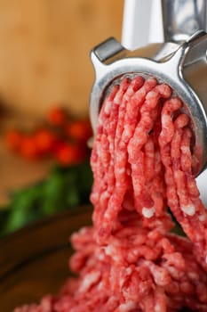 Concept of healthy and proper nutrition, cooking.Close-up of red ground beef with fat streaks coming out of a metal meat grinder on a wooden background, vegetables and herbs are visible out of focus. High quality photo