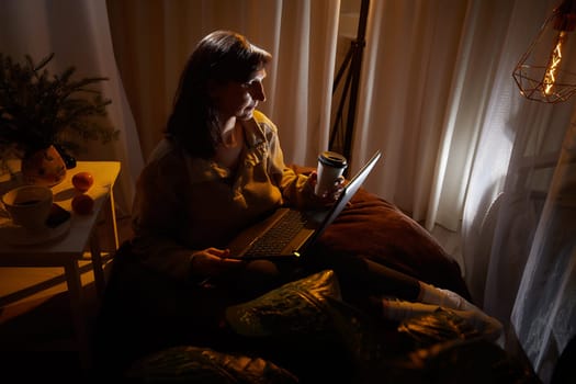 A middle-aged woman sits comfortably in her apartment, savoring coffee while using her laptop during a relaxing evening.