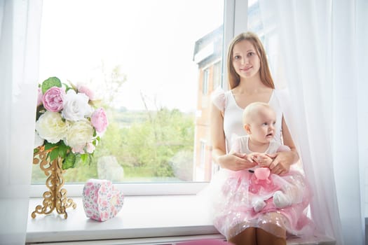 A mother sits with her baby by a bright window, surrounded by flowers and a decorative heart box.