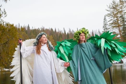 Two girls dressed in flowing white and green attire hold hands by a serene lake, symbolizing the beautiful meeting of winter and spring.