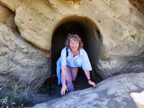A middle-aged woman poses playfully at the entrance of a cave, enjoying her outdoor travel experience.