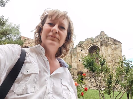 A female travel blogger captures a selfie at a historical site surrounded by lush greenery and vibrant flowers on her journey.